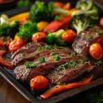 A close-up of a sheet pan filled with sliced flank steak and colorful roasted vegetables including broccoli, bell pepper, and cherry tomatoes.