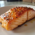 Close-up of a honey glazed salmon fillet on a white plate, garnished with green onions, with soft lighting.
