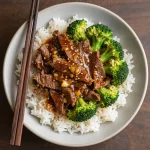 A close-up shot of a serving of Chinese beef and broccoli stir-fry in a white bowl, garnished with sesame seeds.